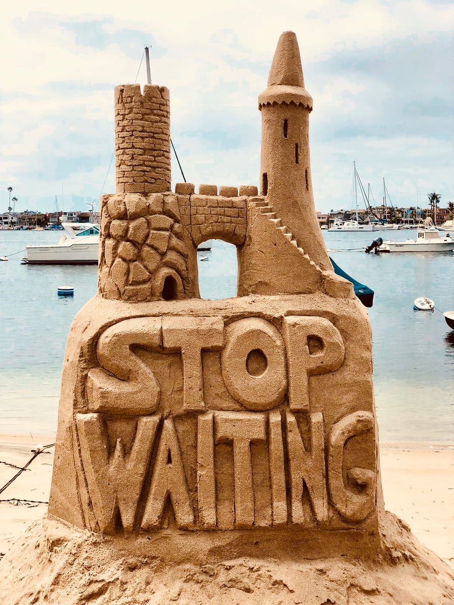 Intricate sand castle with "Stop Waiting" message by sea.