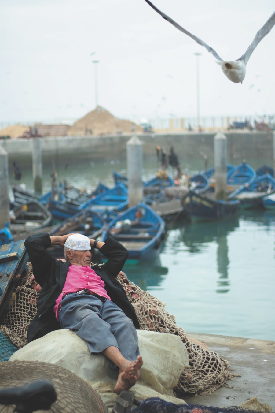 Man relaxing on fishing nets at harbor with boats.