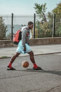 Man dribbling basketball on city street with backpack.