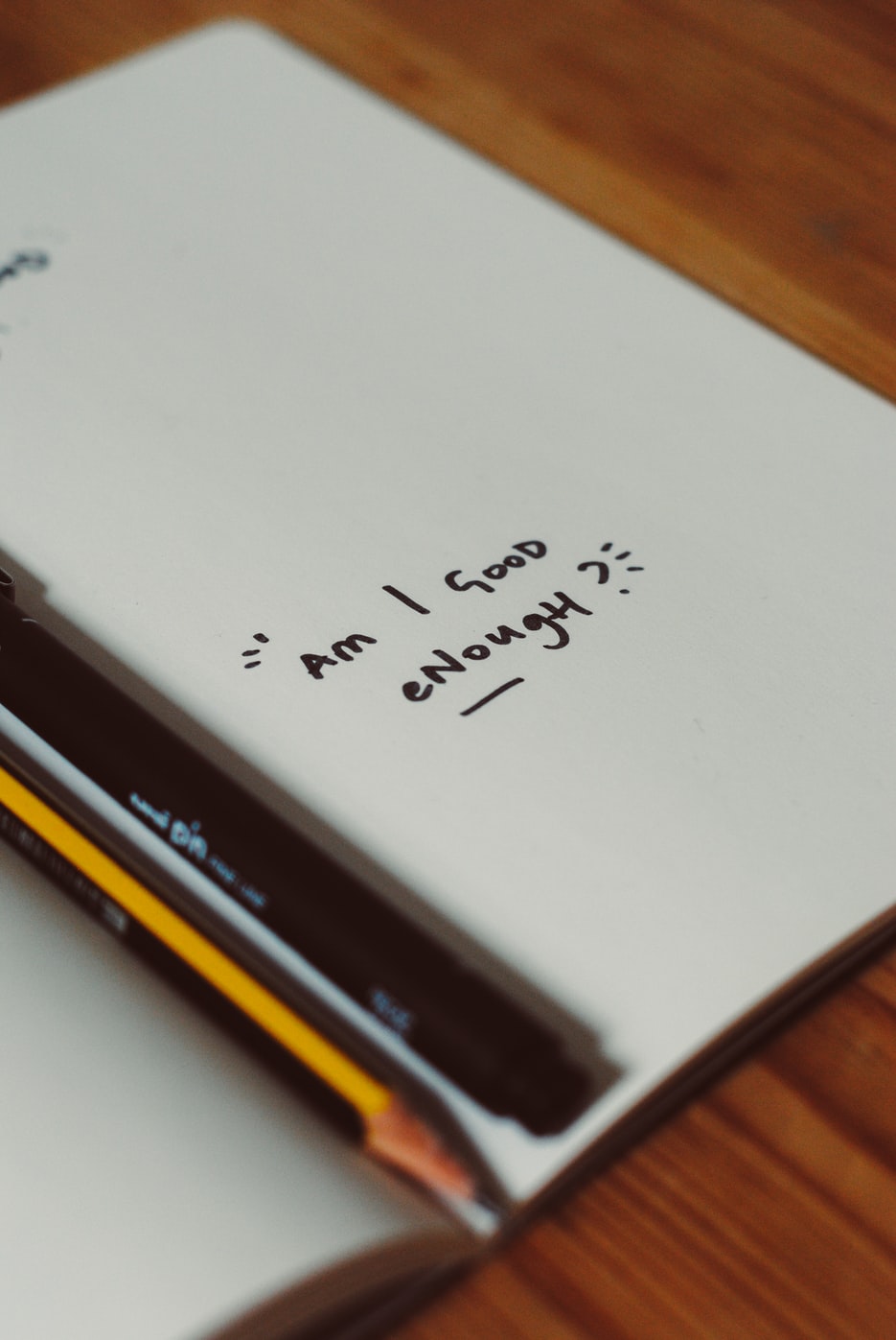 Notebook questioning self-worth with pens on wood desk.