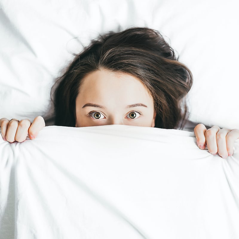 Woman peeking over white bedsheet, eyes open wide.