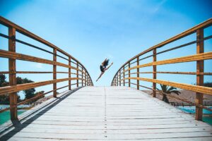 Person jumping on curved wooden bridge under blue sky.