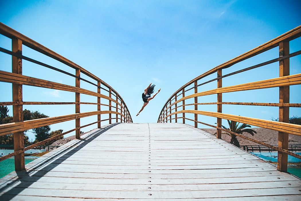 Person jumping on curved wooden bridge under blue sky.